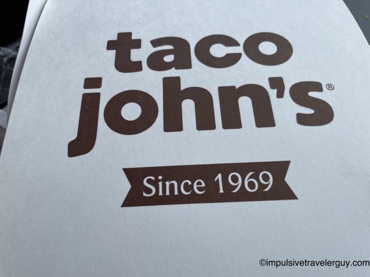 Close-up of a Taco John's takeout box with the brown-on-white logo and 'Since 1969' tagline.