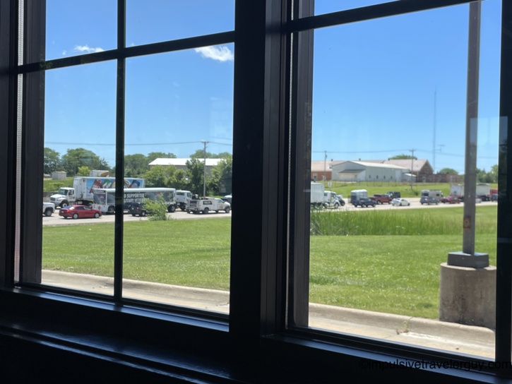 View through a plate-glass window at Taco John's looking out at a commercial lot with delivery trucks, work vehicles, and a 'Proud Supporter of Military Veterans' banner on a parked truck.