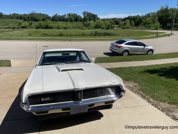 A white 1969 Mercury Cougar Eliminator with Wisconsin collector plates parked in a driveway, with a silver 2007 Ford Focus on the street behind it. Green farmland and a basketball hoop visible across the road on a sunny summer day.