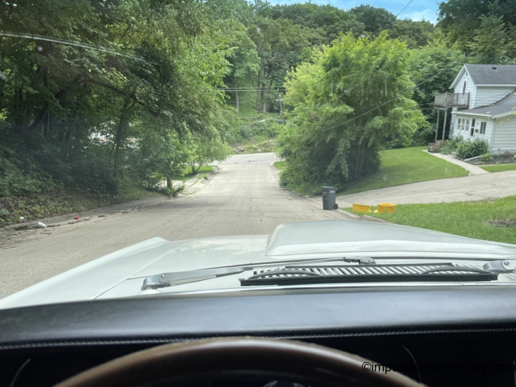Descending a steep, tree-canopied residential street in Dubuque from behind the Cougar's wheel, with old houses perched on the hillside and the road dropping sharply ahead.