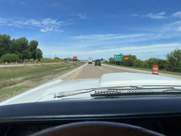 Driving through a construction zone in the Cougar with orange barrels lining both shoulders, a green highway sign ahead pointing north toward Dickeyville and Lancaster as the route heads back into Wisconsin.