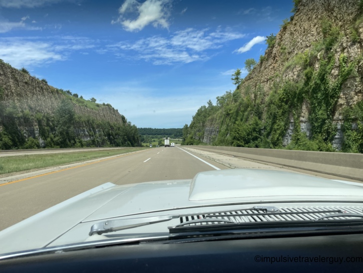 The Cougar passing through a dramatic Driftless Area highway rock cut, exposed limestone bluffs rising on both sides of the divided highway, draped in green vines and vegetation.