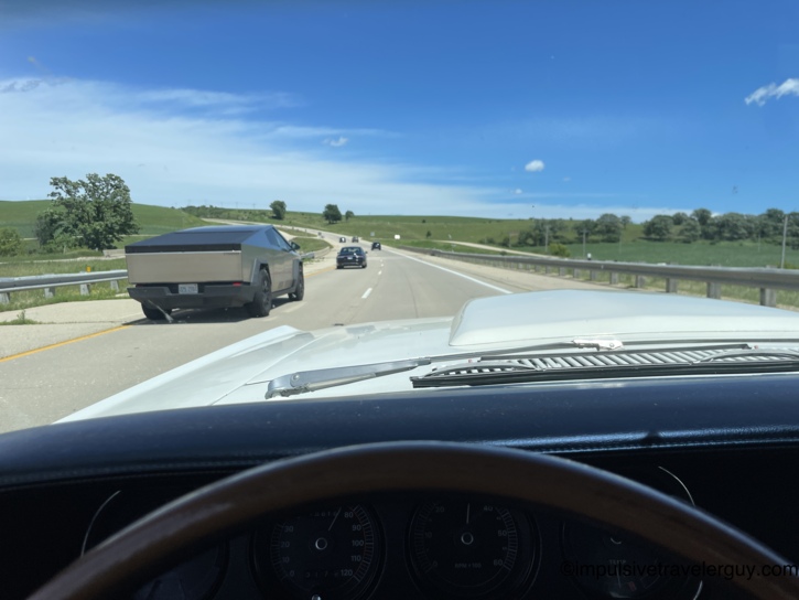 A Tesla Cybertruck passing in the oncoming lane as seen from behind the Cougar's wheel — 1969 meets the future on a rolling green Wisconsin highway.