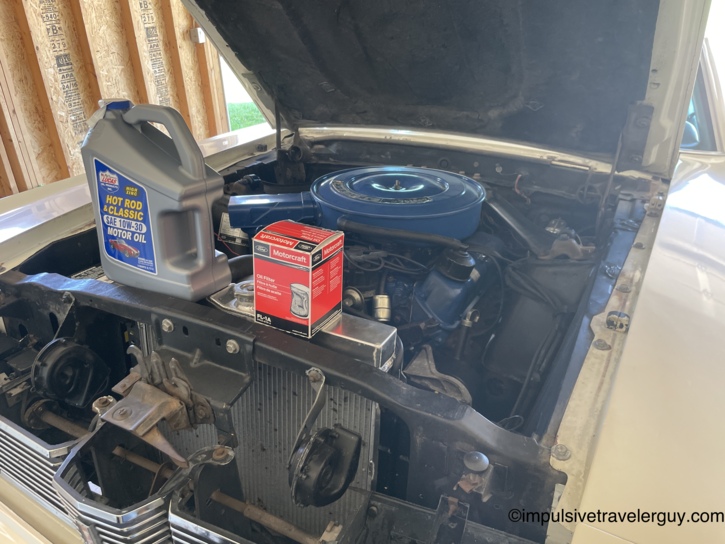Under the Cougar's hood in the garage — a jug of Lucas Hot Rod and Classic SAE 10W-30 high-zinc motor oil and a Motorcraft FL-1A oil filter box sitting on the radiator support next to the Ford-blue air cleaner, ready for an oil change.