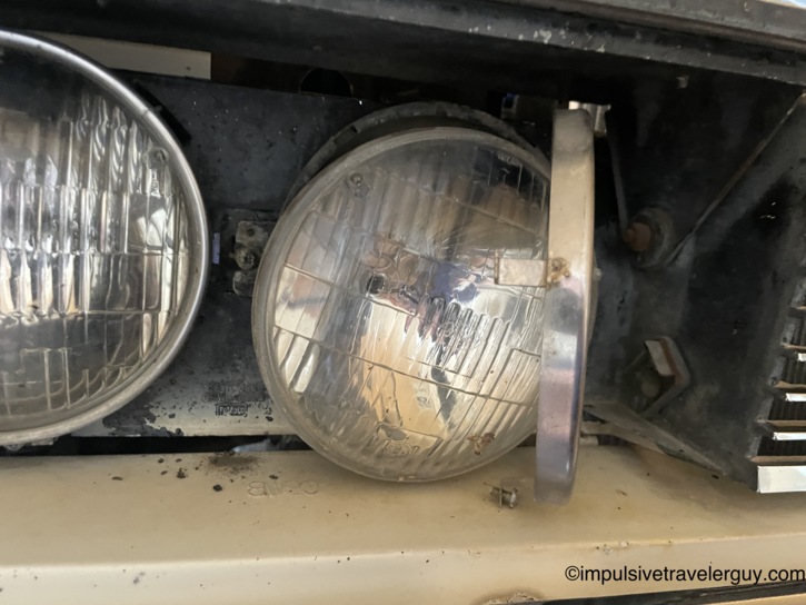Close-up behind the Cougar's open grille showing the dual sealed-beam headlights, with the inner high-beam bulb pulled forward in its retaining ring, ready to be swapped out.