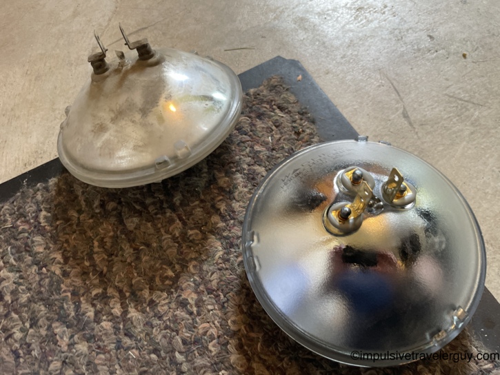 Old versus new sealed-beam headlight bulbs side by side on the garage floor — the original on the left with a cloudy, heat-hazed reflector, the fresh replacement on the right with a clean mirror finish.