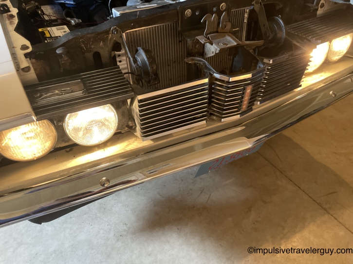 Front of the Cougar in the garage after the headlight swap, all four sealed beams now glowing evenly through the chrome grille, hood still up and blue engine visible behind.