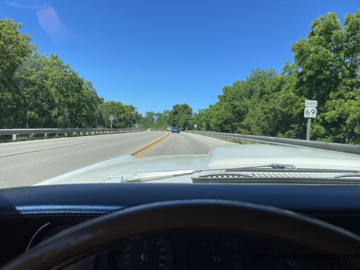 View from behind the wheel of the Cougar cruising south on Highway 69 through a green, tree-lined corridor. The chrome wipers, white hood scoop, and vintage speedometer are visible across the bottom of the frame.