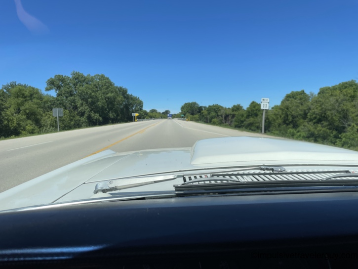 Driving west on Highway 11 from the Cougar's interior, open two-lane road stretching flat through summer-green countryside under a clear blue sky.