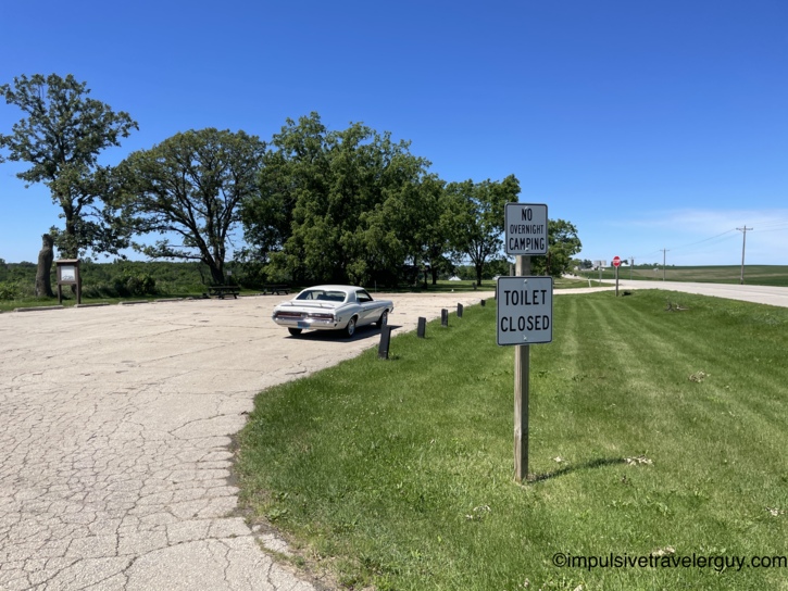 The white Cougar parked at a weathered roadside rest area with cracked pavement, mature shade trees, and signs reading 'No Overnight Camping' and 'Toilet Closed.'
