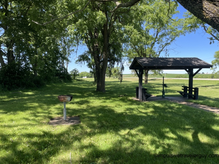 A shaded picnic shelter with a hand-pump well and a pedestal charcoal grill under large trees at the rest area, rolling green farmland visible in the background.