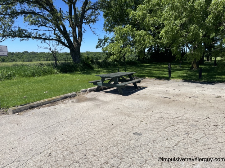 A lone green picnic table sitting on a cracked concrete pad at the rest area, with a large oak tree and open pasture behind it.
