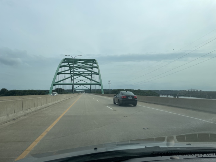 Crossing a large green steel arch bridge over a wide river on a multi-lane highway under cloudy skies.