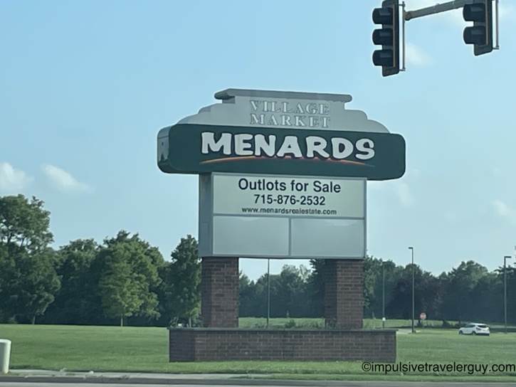 Menards Village Market roadside sign advertising outlots for sale along a tree-lined rural highway.