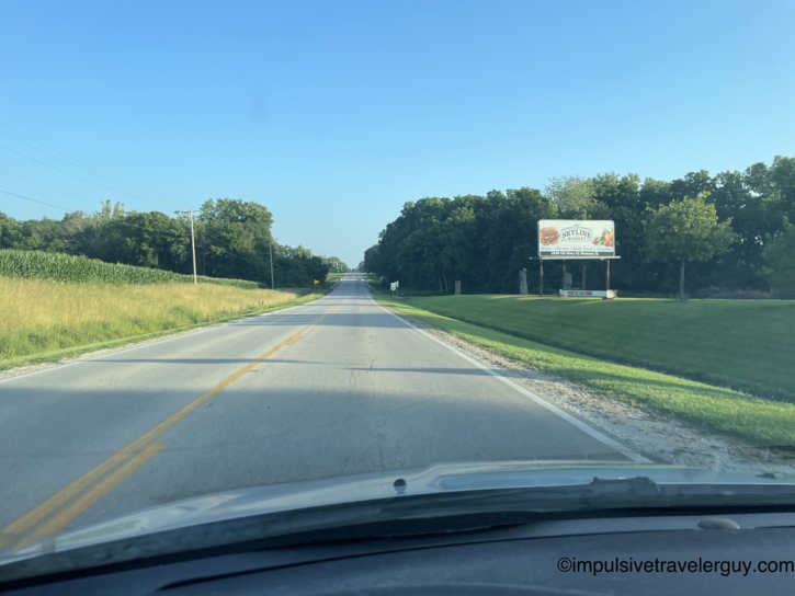 Rural two-lane highway stretching straight ahead through cornfields with a Skyline Market billboard on the right.