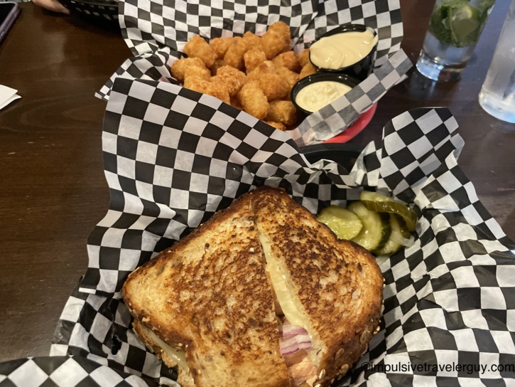 Salmon grilled cheese sandwich on multigrain bread served with tater tots, pickles, and dipping sauces in checkered paper-lined baskets.