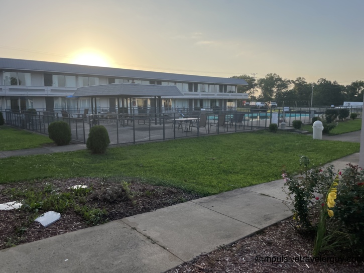 Two-story motel courtyard with fenced outdoor pool area, patio furniture, and landscaped flower beds at golden hour.