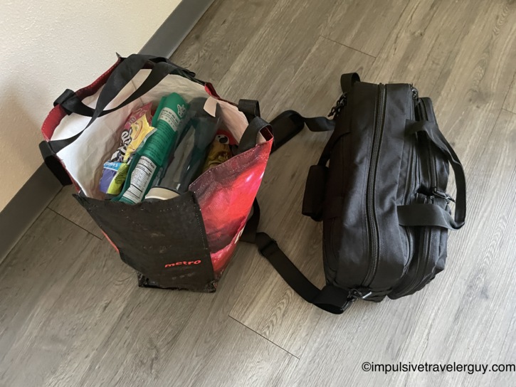 Reusable Metro shopping bag stuffed with road trip snacks and toiletries next to a black travel bag on the motel room floor.