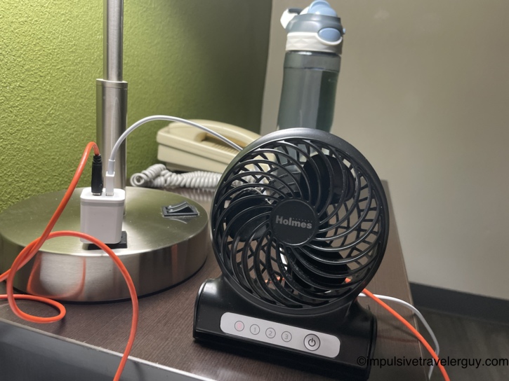 Small black Holmes personal fan plugged in on the motel nightstand next to a lamp, phone charger cable, and water bottle.