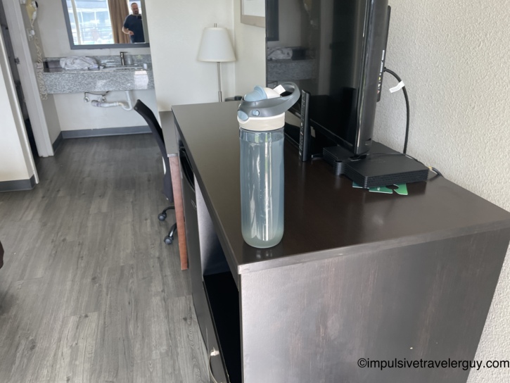 Motel room interior from the dresser side showing water bottle, flat-screen TV, desk area, and open bathroom vanity in the background.