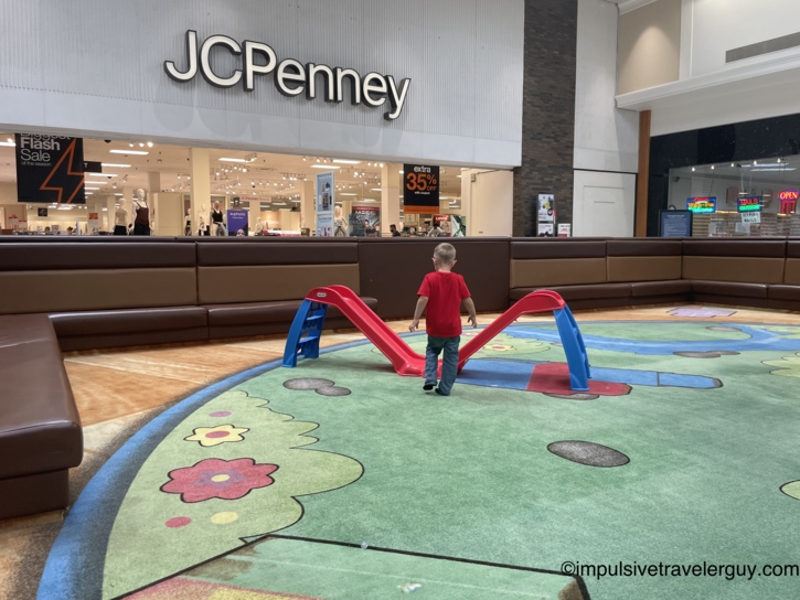 Small child in a red shirt exploring the play area slide near JCPenney at an indoor shopping mall.