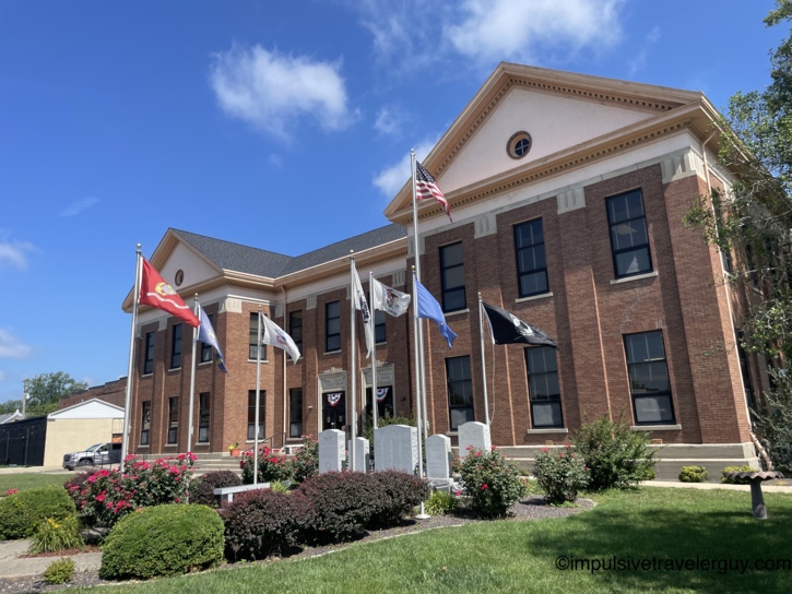 Red brick county courthouse with classical pediment, round window, military service flags, veterans memorial stones, and rose bushes on the front lawn.