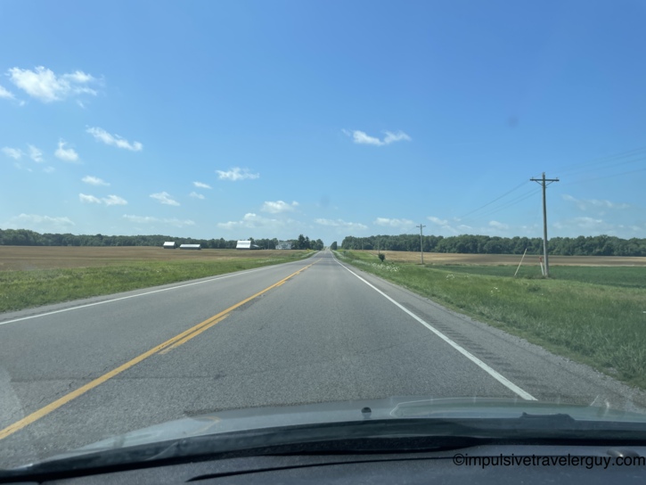 Straight two-lane highway stretching through flat Illinois farmland with white barn structures and power lines under blue sky.