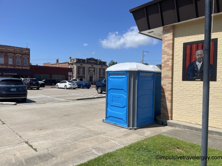 Small-town downtown scene with a blue portable toilet on the sidewalk, a painted portrait mural on a building wall, taproom signage, and historic brick storefronts across the parking lot.