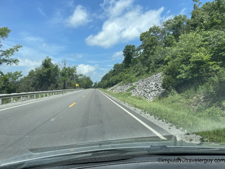 Winding two-lane highway cutting through hilly wooded terrain with a rock pile on the roadside embankment and metal guardrail.
