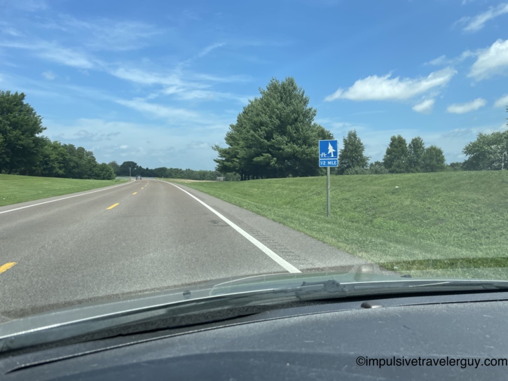 Rural highway with a blue rest area and picnic sign indicating half a mile ahead, rolling green hills and scattered trees under summer sky.