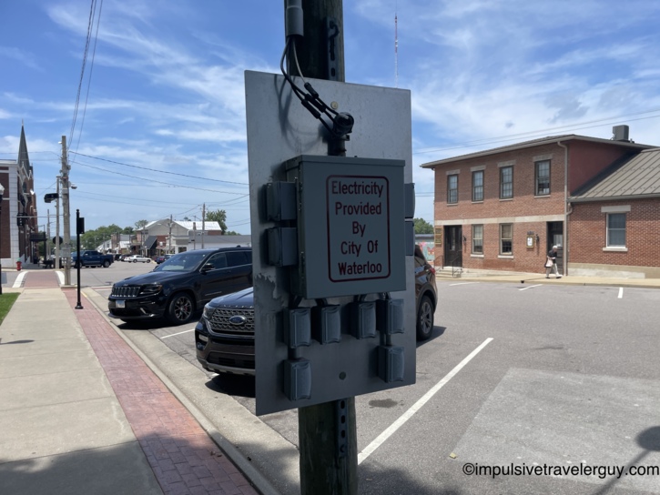 Electrical utility box on a pole in downtown Waterloo, Illinois reading Electricity Provided By City Of Waterloo with a church steeple and brick storefronts visible along the street.
