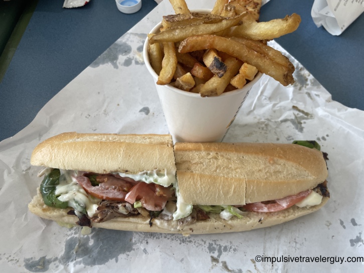 Loaded sub sandwich with steak, peppers, tomatoes, lettuce, and melted cheese alongside a cup of fresh-cut fries.