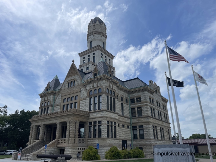 Jersey County Courthouse, an ornate Richardsonian Romanesque stone building with a tall clock tower, copper-patina turrets, and a veterans memorial with American and Illinois flags on the lawn.