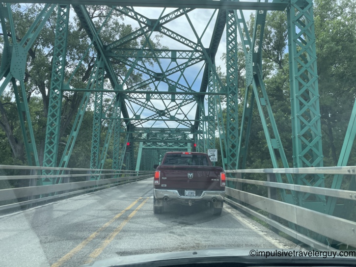 Following a Ram pickup across a narrow single-lane green steel truss bridge with a Stop Here On Red sign for alternating traffic.