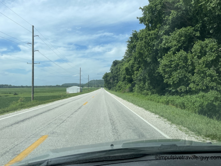 Two-lane rural highway running straight through Illinois River valley bottomland with a metal barn, power lines, dense tree line on the right, and bluffs in the distance.