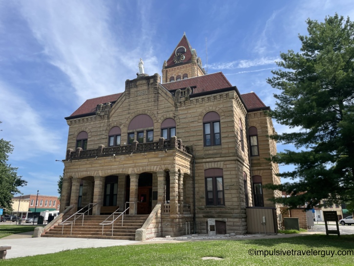 Romanesque sandstone courthouse dated 1891 with red tile roof, clock tower, arched entryway with stone columns, and a statue atop the central gable on a town square lawn.