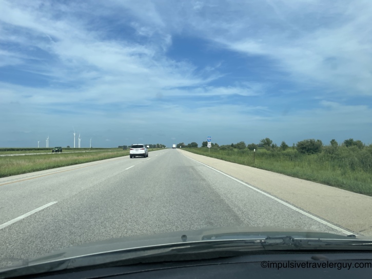 Driving on Interstate 72 and US Route 36 through flat central Illinois with wind turbines visible on the left horizon under wispy clouds.