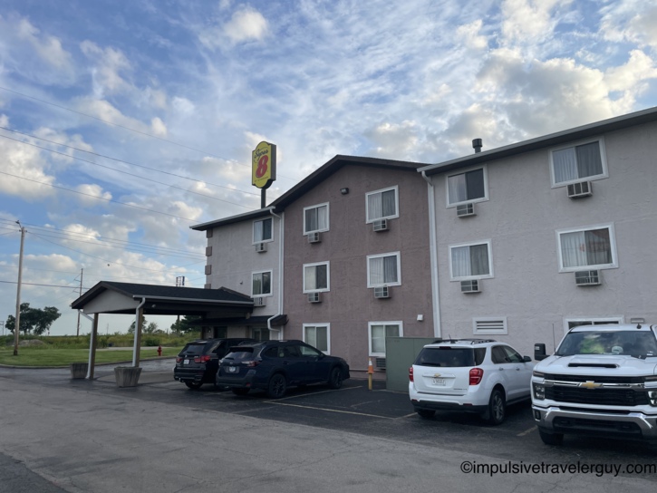 Super 8 motel exterior, a three-story stucco building with the yellow-and-red sign, covered entrance portico, and vehicles in the parking lot under partly cloudy skies.