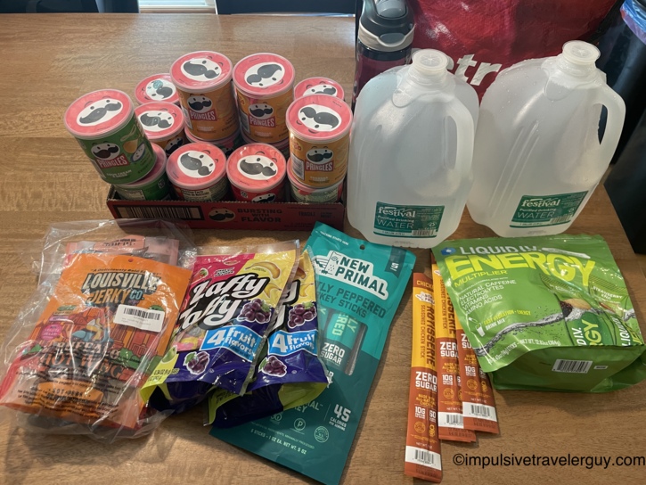 Spread of road trip snacks on a table including a case of Pringles, gallon water jugs, Louisville Jerky, Laffy Taffy, New Primal turkey sticks, chicken sticks, and Liquid IV Energy packets.