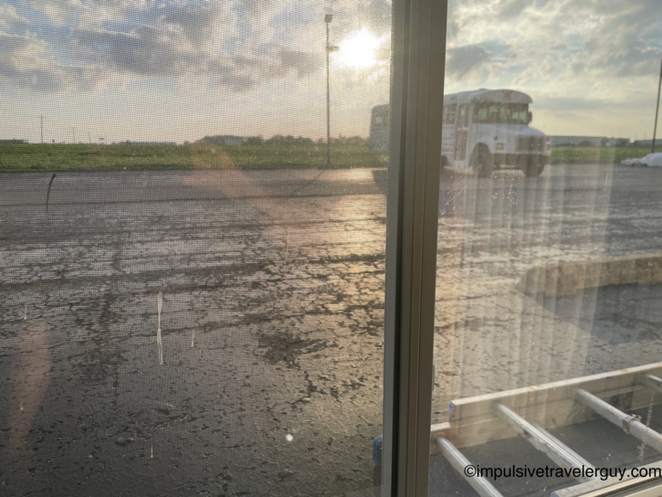 View through a motel window screen at sunrise showing a cracked parking lot with a school bus parked in the background.