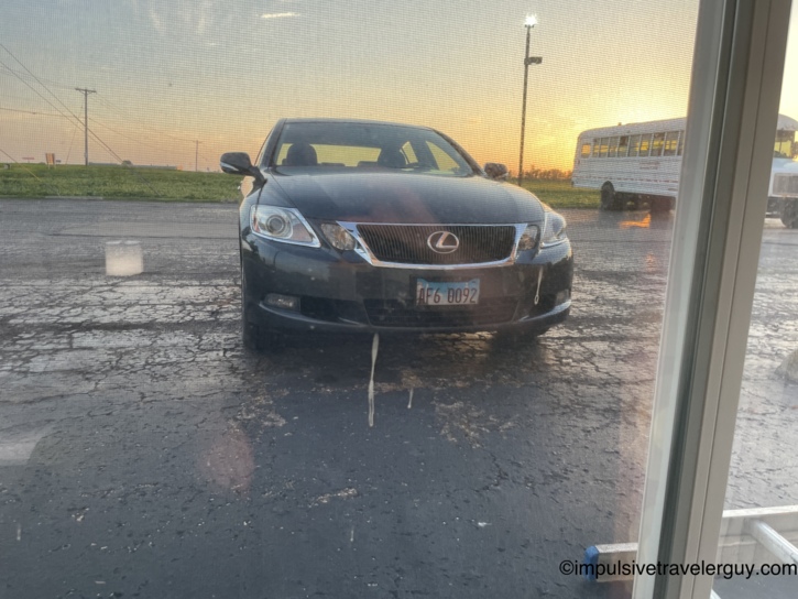 Black Lexus sedan parked directly outside the motel room window at sunset with a school bus visible behind it.
