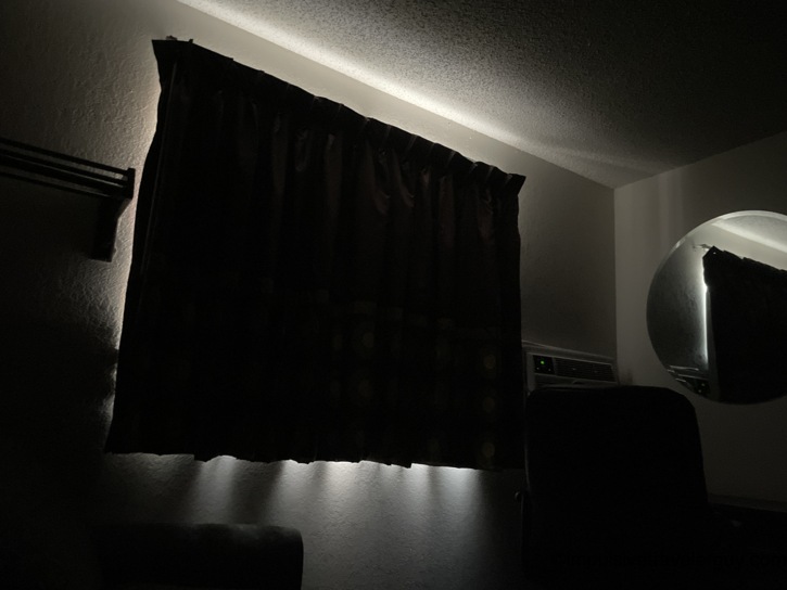 Dark motel room at night with blackout curtains drawn, light leaking around the edges, and the green glow of the AC unit indicator visible.