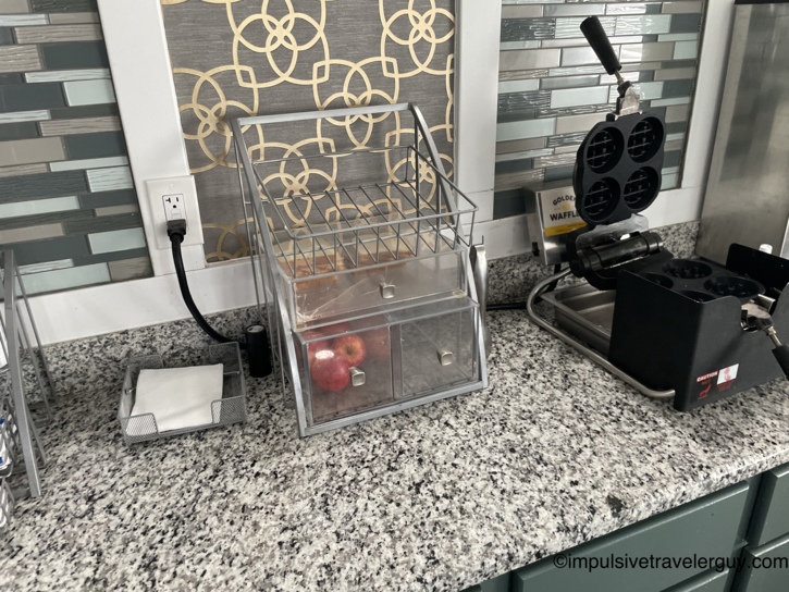 Budget motel continental breakfast station with a pastry and fruit display case holding an apple and baked goods, next to a Golden Waffle maker on a granite counter with mosaic tile backsplash.
