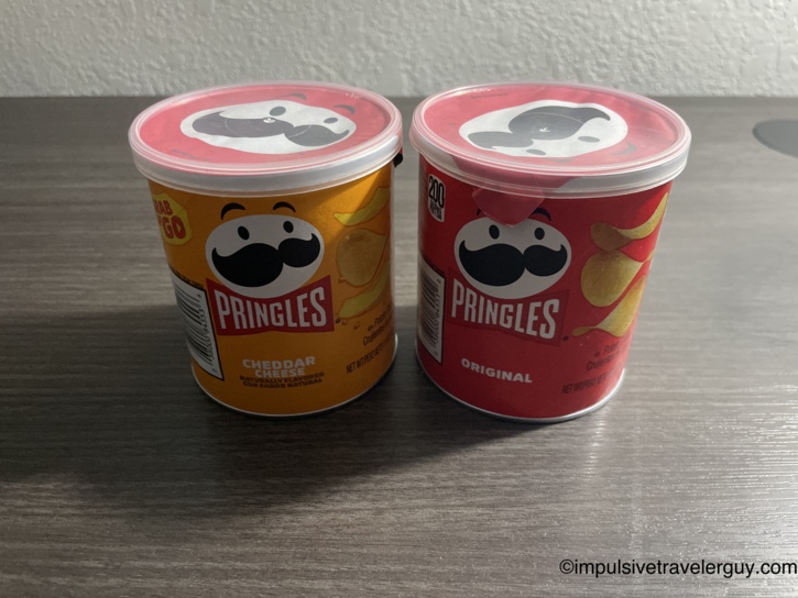 Two grab-and-go Pringles cans on the motel room dresser, Cheddar Cheese and Original flavors.
