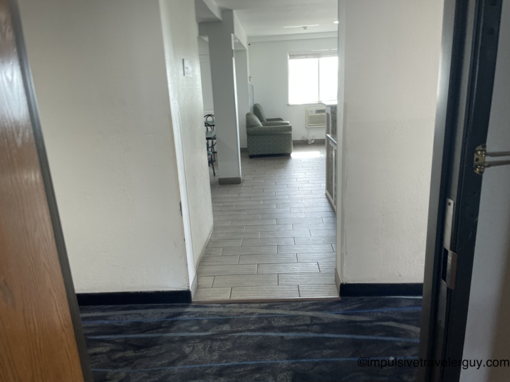 Motel lobby hallway looking into a small sitting area with a green upholstered chair, tile flooring, window AC unit, and carpeted corridor.