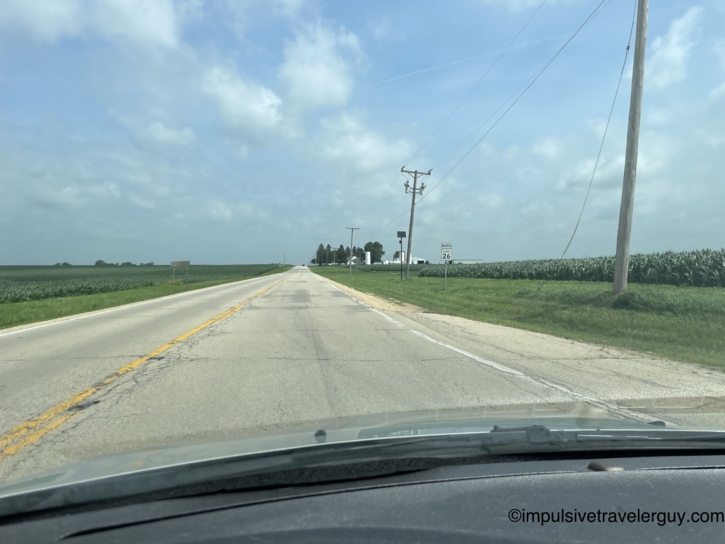 Driving north on Illinois Route 26 through open farmland with corn on both sides, a farm with silos and barns ahead, and partly cloudy skies.