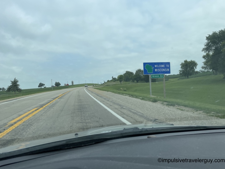 Welcome to Wisconsin sign at the Green County state line on a rural highway with rolling green hills and a red barn with silo visible on the left.