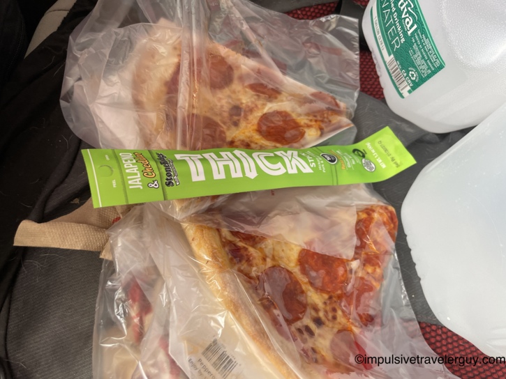 Pepperoni pizza slices in plastic bags and a Stonefire Jalapeno and Cheddar Thick Stick on the car seat next to a gallon jug of Festival water.