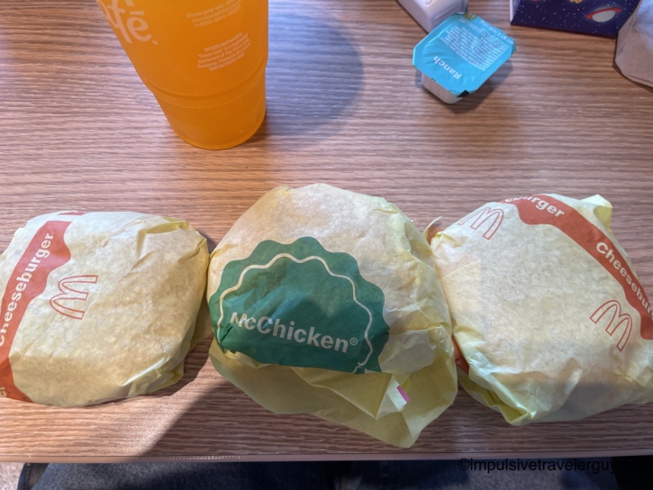 Three wrapped McDonald's sandwiches resting on a wooden table alongside an orange drink cup and a packet of ranch dressing from the lunch stop.