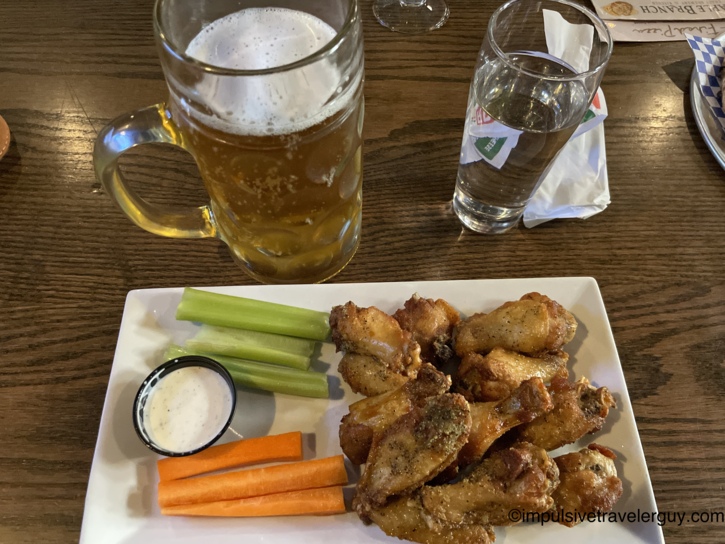A large glass mug of beer sitting on a wooden table next to a plate filled with seasoned chicken wings.